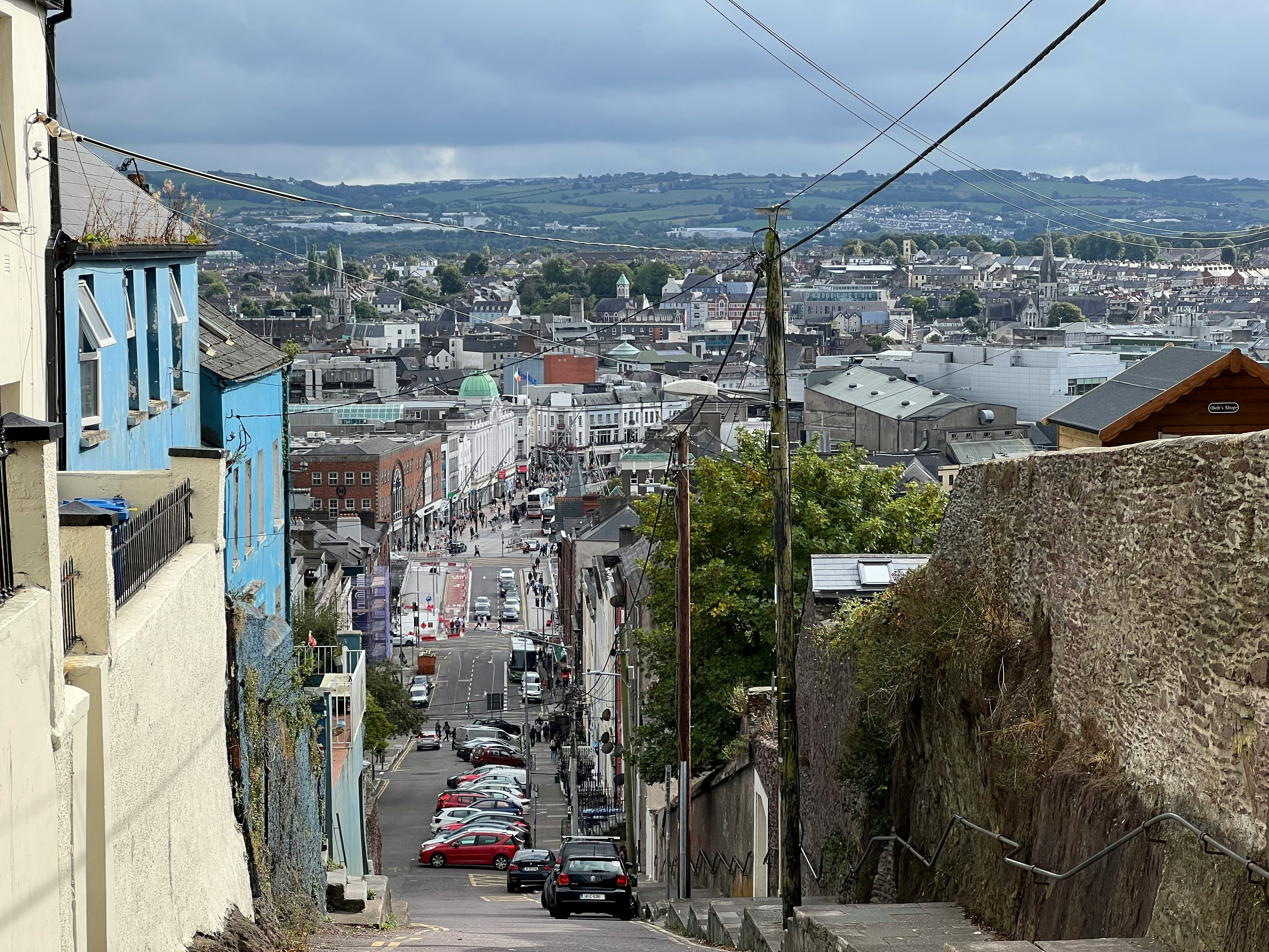 View down a steep Cork street towards the city centre, with rooftops, church spires, hills and overhead wires under a grey sky.